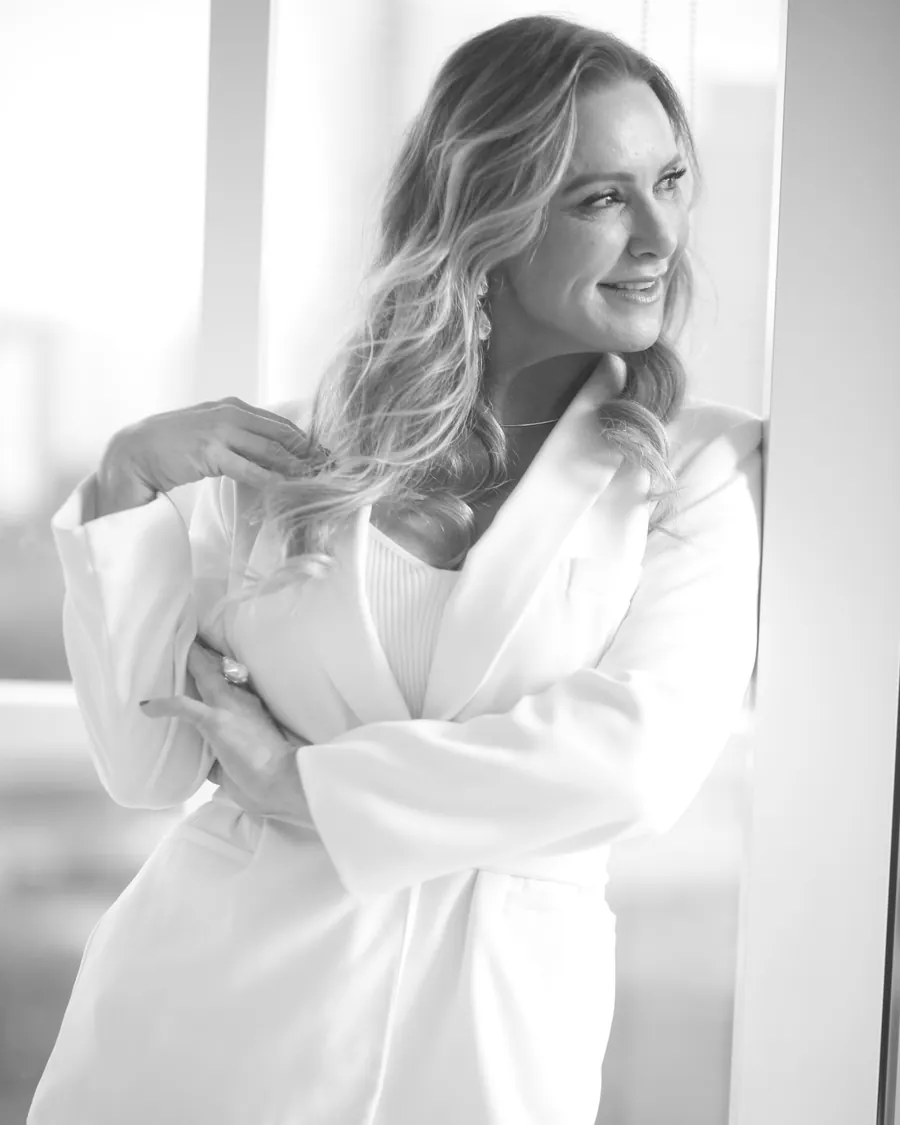 Patrícia Sivieri de terninho branco, sorrindo, junto à janela
